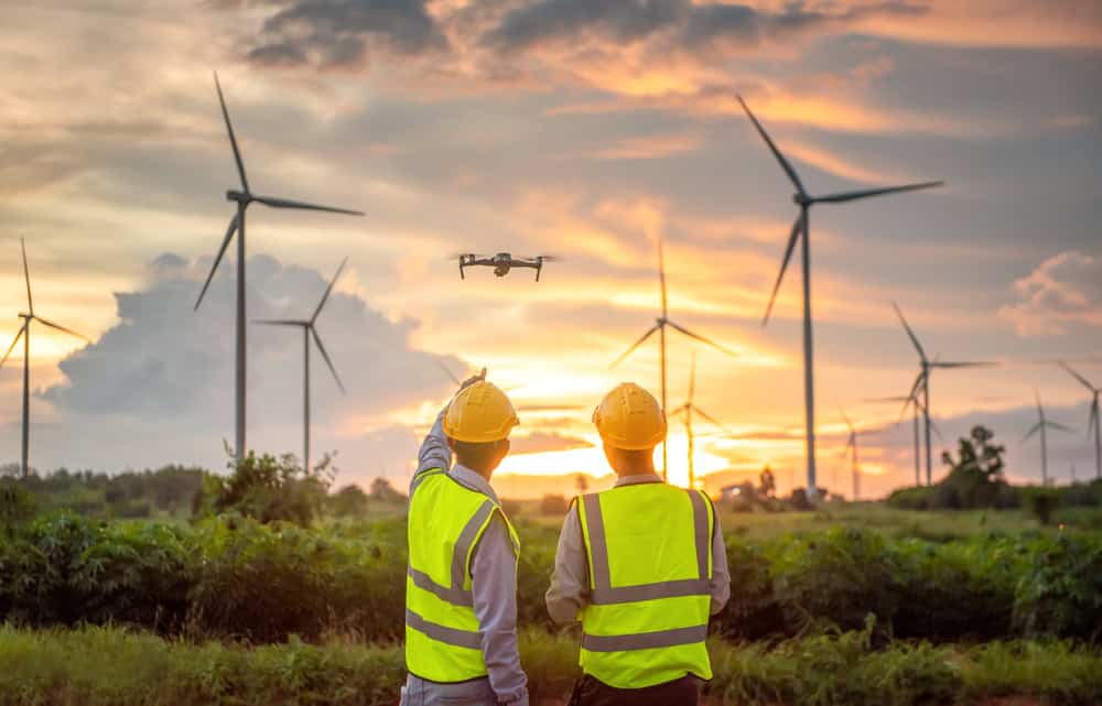 Two workers in high-visibility vests and helmets, likely land surveyors from Martin County, observe a drone flying near a wind farm at sunset. Wind turbines stand tall against a colorful sky, blending orange, pink, and blue hues as the sun sets in the background.