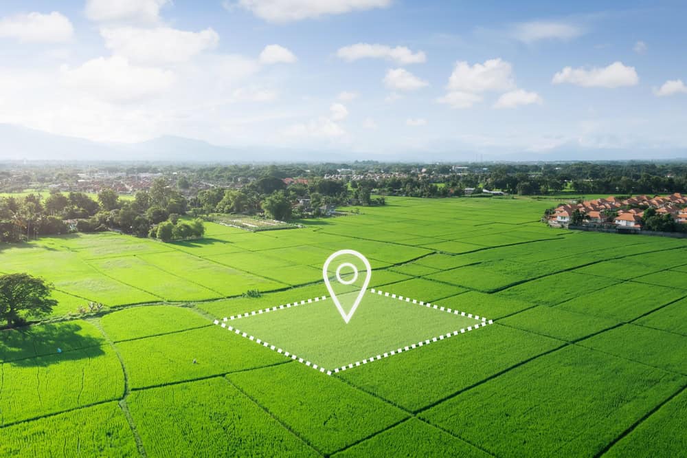 Aerial view of a vast, lush green field under a clear blue sky. A white location pin icon marks a dotted rectangular area within the field, surveyed by Land Surveyors Martin County. Houses and mountains are visible in the distant background.