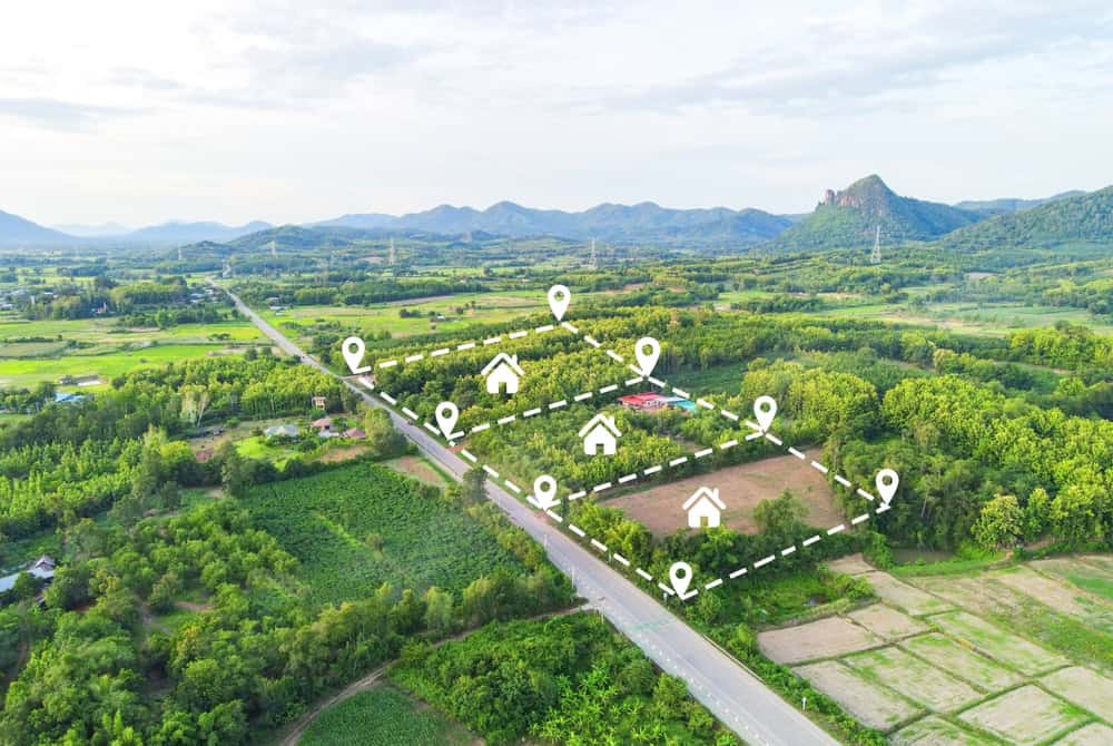 Aerial view of a rural landscape in Indian River County with green fields and scattered trees. A road runs through the scene, where digital icons mark plots within dashed lines, ideal for land surveyors. Hills and distant mountains provide a stunning backdrop.