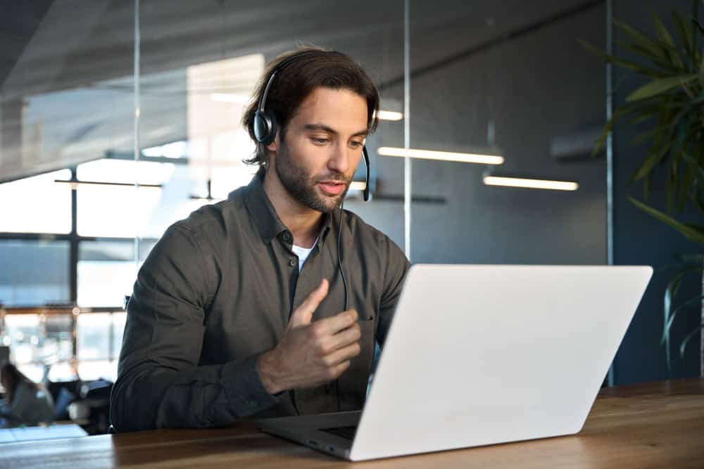 A man with long hair and a beard is sitting at a table wearing a headset, looking at a laptop. He appears engaged, possibly discussing projects with land surveyors from Okeechobee County. The modern office setting features large windows and ambient lighting.