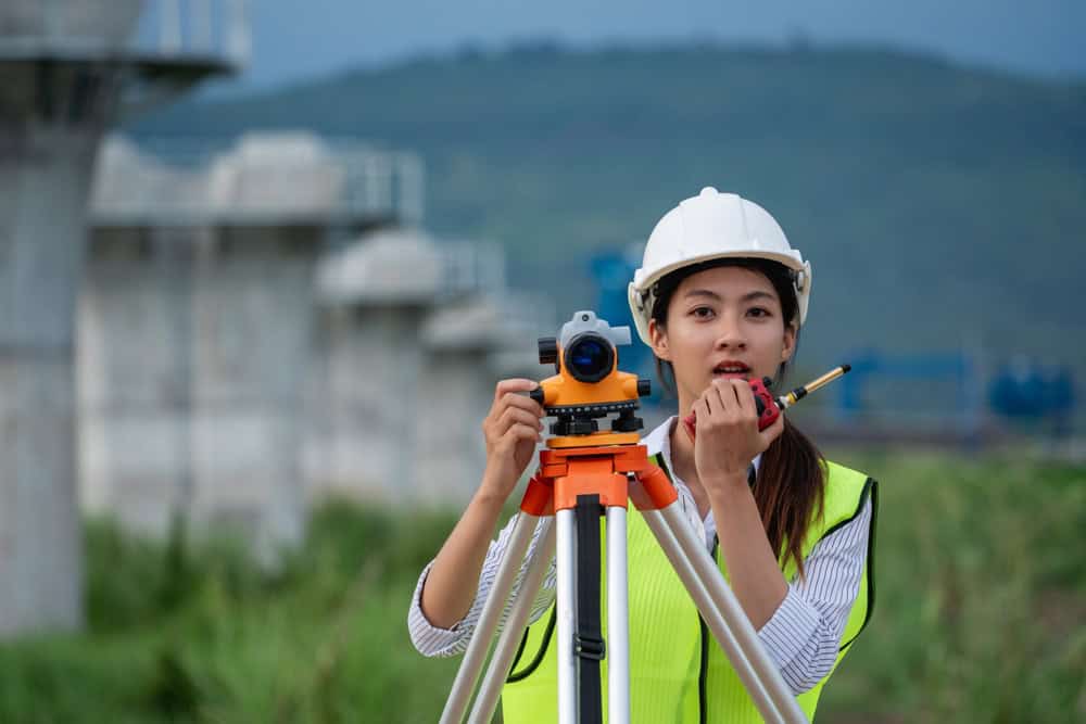 A woman in a hard hat and safety vest uses surveying equipment outdoors, representing the skilled land surveyors of Indian River County. She holds a radio amidst the grassy area, with construction structures and hills serving as her backdrop.