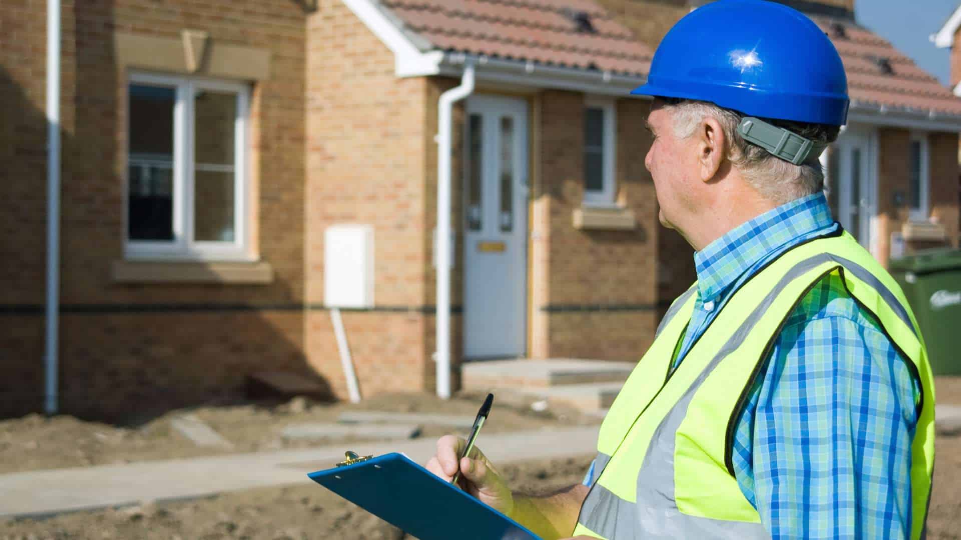 A construction worker in a blue hard hat and reflective vest holds a clipboard and pen, meticulously inspecting the exterior of a brick house. The scene, reminiscent of land surveyors in Indian River County, suggests a keen focus on building inspection or site management.