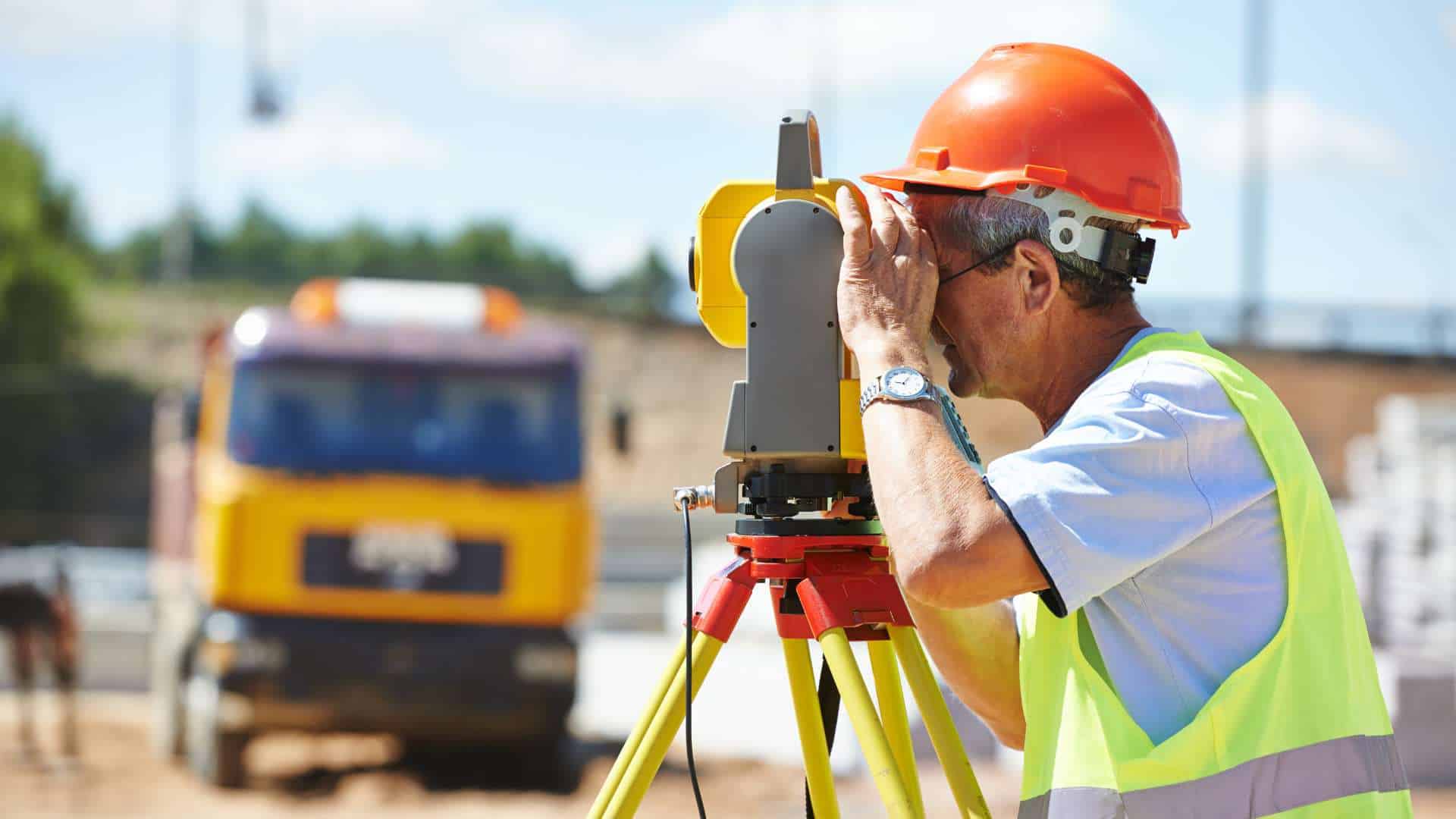 A construction worker in a high-visibility vest and orange helmet expertly uses a theodolite on a tripod at the bustling construction site, embodying the precision of Land Surveyors in Indian River County. A blurred yellow truck looms in the background, symbolizing progress and development.