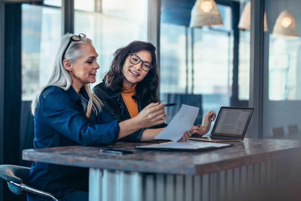 Two women sit at a desk, engrossed in reviewing documents. One holds a pen while the other smiles, their laptop open as they delve into details. Their discussion seems focused and professional, much like land surveyors in Martin County collaborating on an important project.