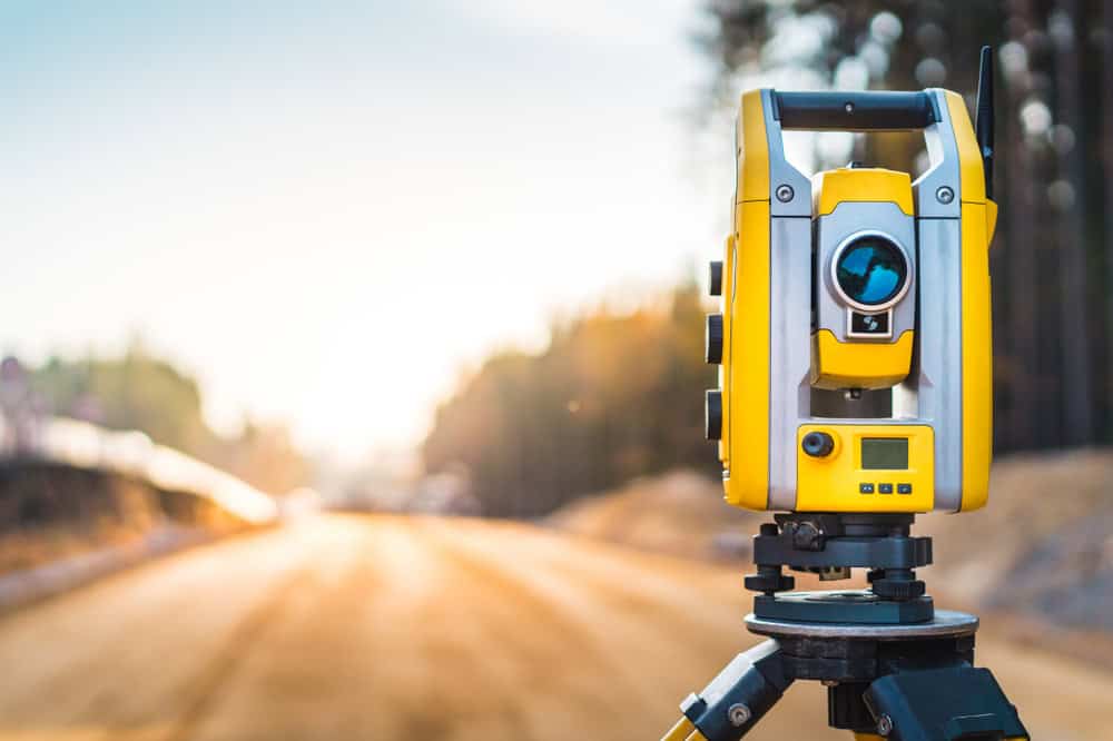 A yellow surveying total station, essential for Land Surveyors in Martin County, stands on a tripod focusing on a blurred construction site. A dirt road stretches under the bright sky, framed by forested edges that hint at early morning or late afternoon light.