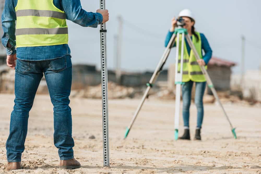 Two construction workers wearing yellow safety vests and jeans are on a sandy construction site. One holds a leveling rod, while the other uses a theodolite on a tripod. Under a clear sky, these land surveyors bring precision to their task in Indian River County.