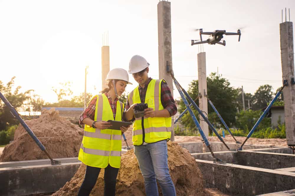 Two construction workers in safety vests and helmets examine a tablet and remote control as their drone hovers nearby at the site. With concrete pillars, piles of dirt, trees, and a sunset sky as their backdrop, the scene unfolds like those commonly managed by Land Surveyors in Martin County.