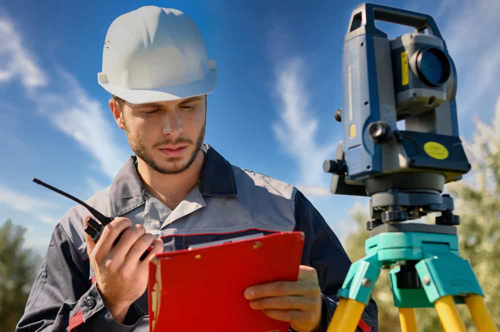 A construction worker in a hard hat uses a walkie-talkie and checks notes on a clipboard as he assists Land Surveyors Martin County. A surveying instrument is set up on a tripod nearby, under a clear sky with some clouds in the background.