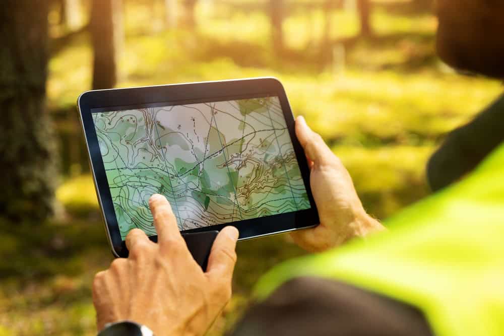 A person wearing a bright yellow vest, likely one of the Land Surveyors in Martin County, is outdoors holding a tablet displaying a topographic map. The background reveals a sunlit, grassy area with trees.