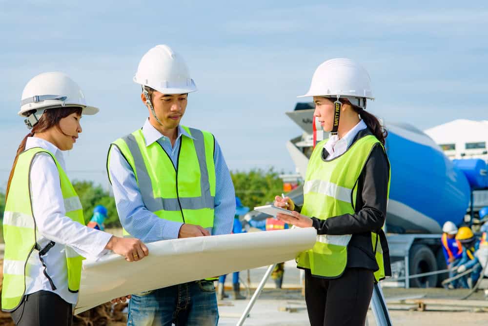 Three construction workers in safety helmets and vests stand at a site, reviewing plans, with a cement mixer in the background. One worker, possibly a land surveyor from Indian River County, holds a clipboard while another holds a large blueprint.