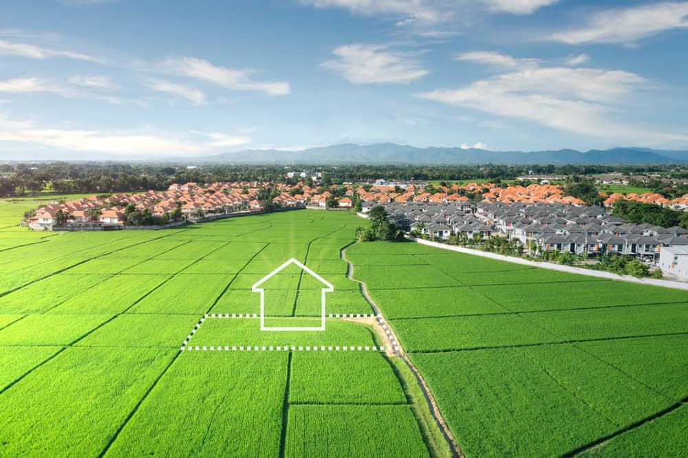 In the foreground, a large, green open field with an outline of a house drawn on it symbolizes precision in design. In the background, there’s a picturesque suburban neighborhood against mountainous horizons. Expertise from Land Surveyors Indian River County is evident in every measured detail.