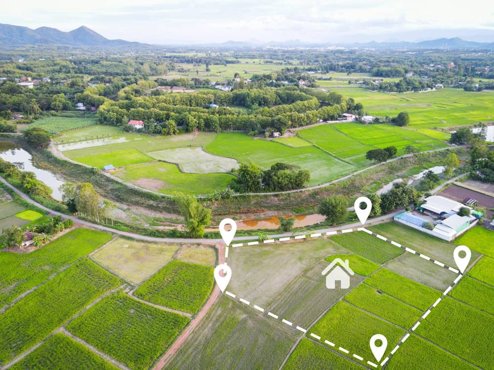 Aerial view of a lush, green agricultural landscape with various fields and buildings. A white dotted boundary highlights a plot, marked by Land Surveyors Martin County, featuring a house icon in the center. The background reveals hills and distant mountains.