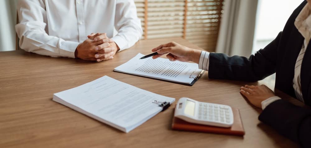 Two people sit across from each other at a table, with one pointing a pen at a document on a clipboard. A calculator and notepad lie nearby, hinting at a business meeting or interview. The setting could involve land surveyors in Indian River County discussing project details.