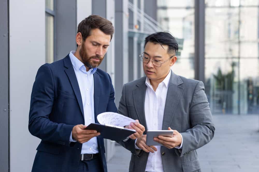 Two businessmen in suits stand outside an office building. One man reviews documents on a clipboard, while the other holds a tablet. They discuss details akin to land surveyors in Martin County, all set against a backdrop of modern architecture.