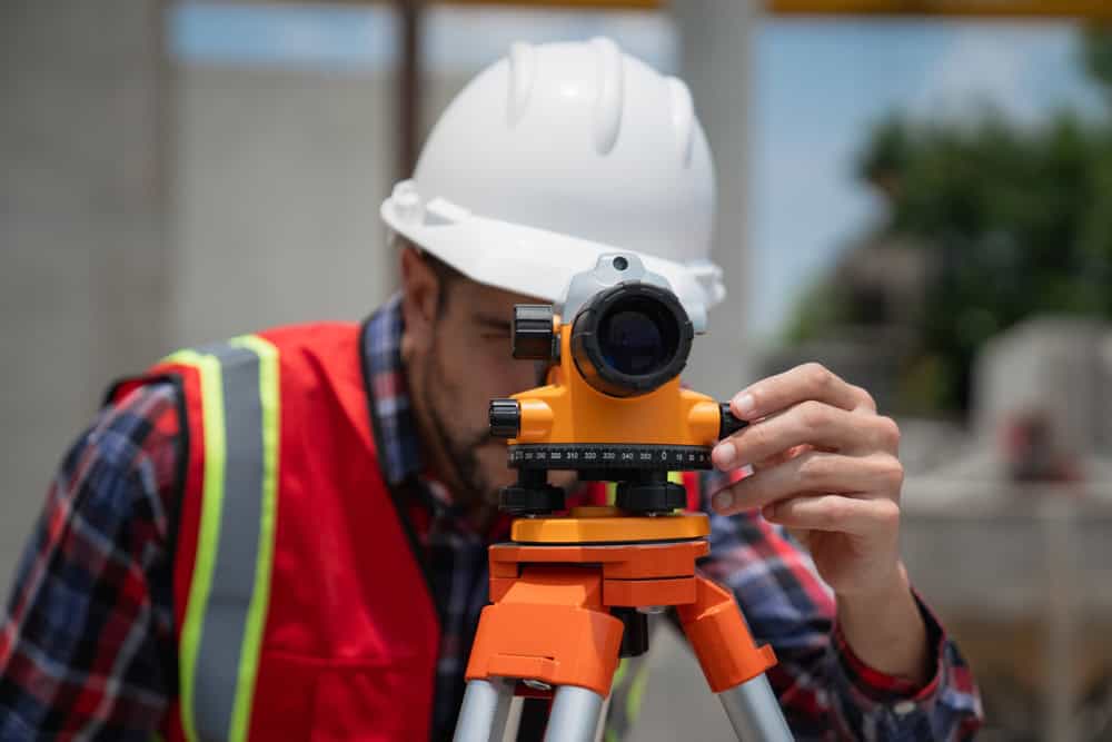 A construction worker wearing a white hard hat and red safety vest uses a theodolite on a site. The background is blurred, highlighting his precise work with the instrument, similar to the experts from Land Surveyors Martin County who ensure accuracy and reliability in every project.