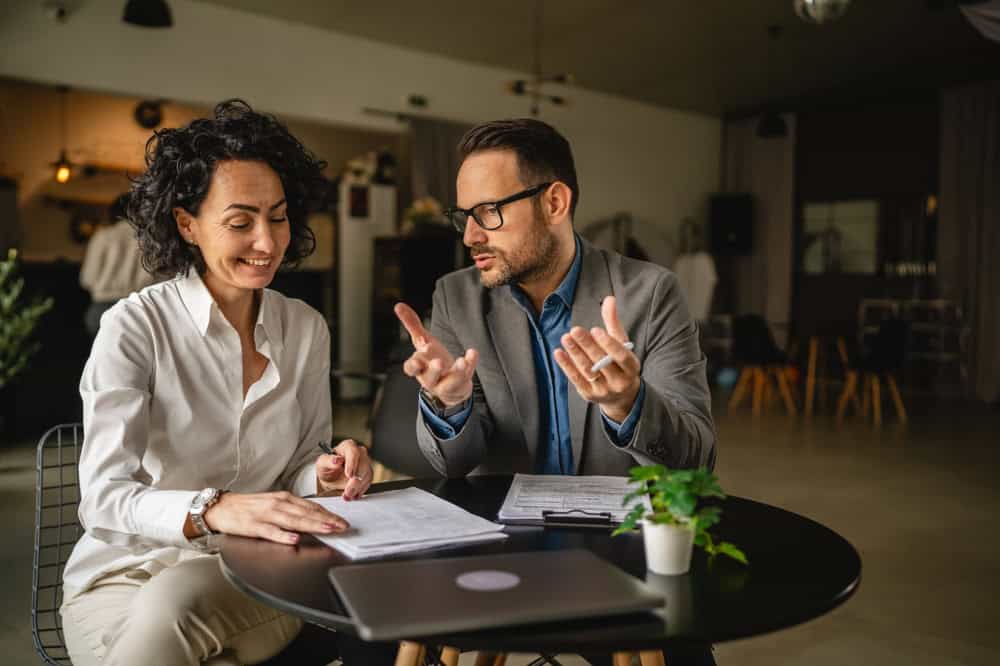 A man and a woman, dressed in formal attire, are sitting at a round table engaged in a discussion. Papers and a laptop share the space with a small potted plant. They appear to be land surveyors from Martin County in a modern, well-lit office setting.