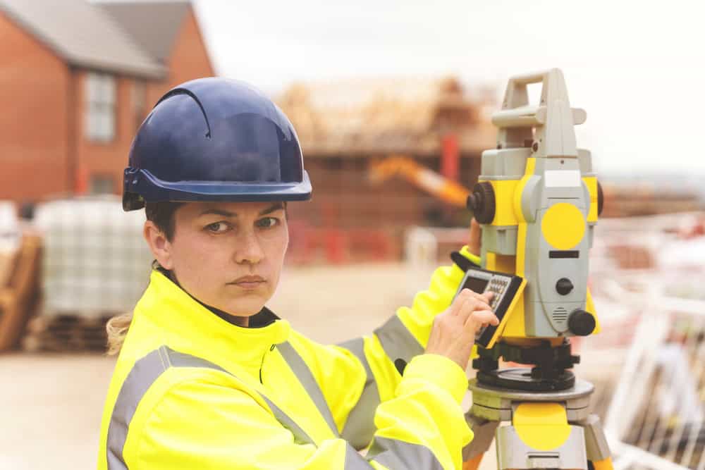 A person wearing a blue hard hat and high-visibility yellow jacket operates a theodolite at a bustling construction site. With partially constructed buildings and materials around, this scene is typical for land surveyors in Martin County.