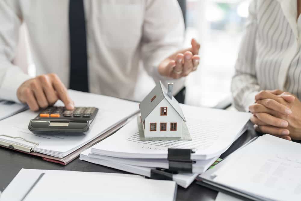 A person in a suit gestures while sitting at a table with papers, a small house model, and a calculator, suggesting a discussion about real estate or finance. Perhaps they're consulting Land Surveyors in Martin County as another individual sits beside them with hands clasped.