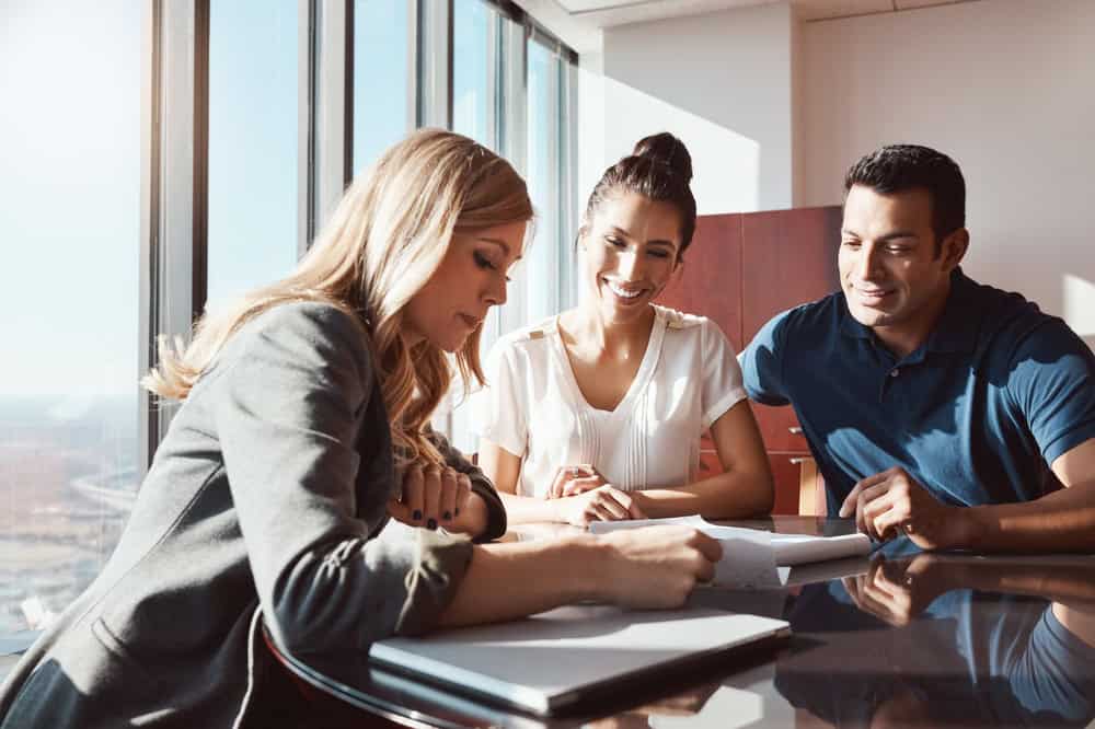 Three people are seated at a table in a bright office. A woman on the left is writing on a document while an experienced land surveyor from Indian River County looks on, smiling, alongside another colleague. A window offers a view of a city skyline in the background.