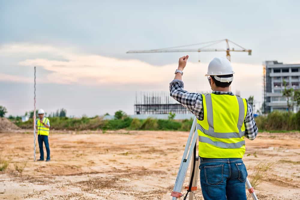 Two land surveyors in reflective vests and hard hats work diligently on a construction site in Martin County. One uses a theodolite while the other holds a measuring pole. A crane and partially constructed building can be seen against the partly cloudy sky above.