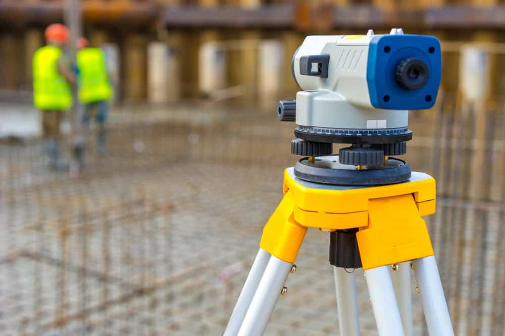 A theodolite on a tripod is set up at a construction site, where Land Surveyors from Indian River County work diligently. Two workers in yellow safety vests and helmets are visible in the blurred background. Rebar and concrete formwork are seen around the site.