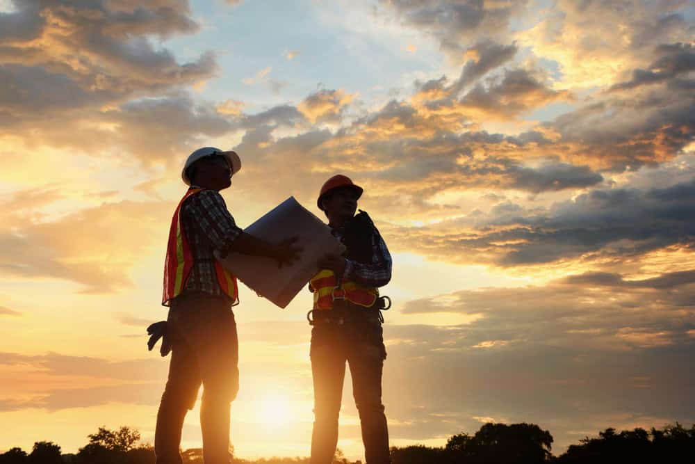 Silhouetted image of two construction workers in safety gear, possibly land surveyors from Indian River County, reviewing blueprints at a construction site during sunset. The sky is dramatic with clouds and warm colors, creating a striking backdrop.
