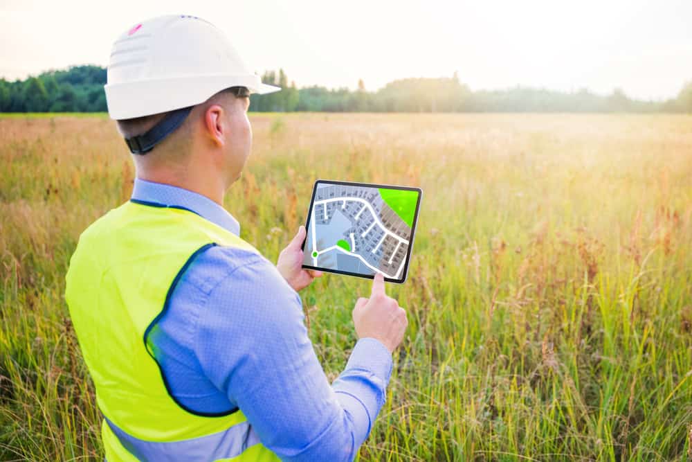 A person in safety gear, including a hard hat and reflective vest, holds a tablet displaying a construction plan while standing in a grassy field under the sunlight, embodying the expertise of land surveyors in Okeechobee County.