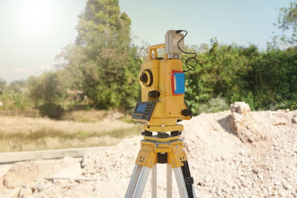 A yellow total station stands on a construction site tripod, bathed in sunlight. In the background, trees and a pile of dirt hint at ongoing surveying work by land surveyors in Okeechobee County, capturing the essence of their meticulous craft in a rural setting.