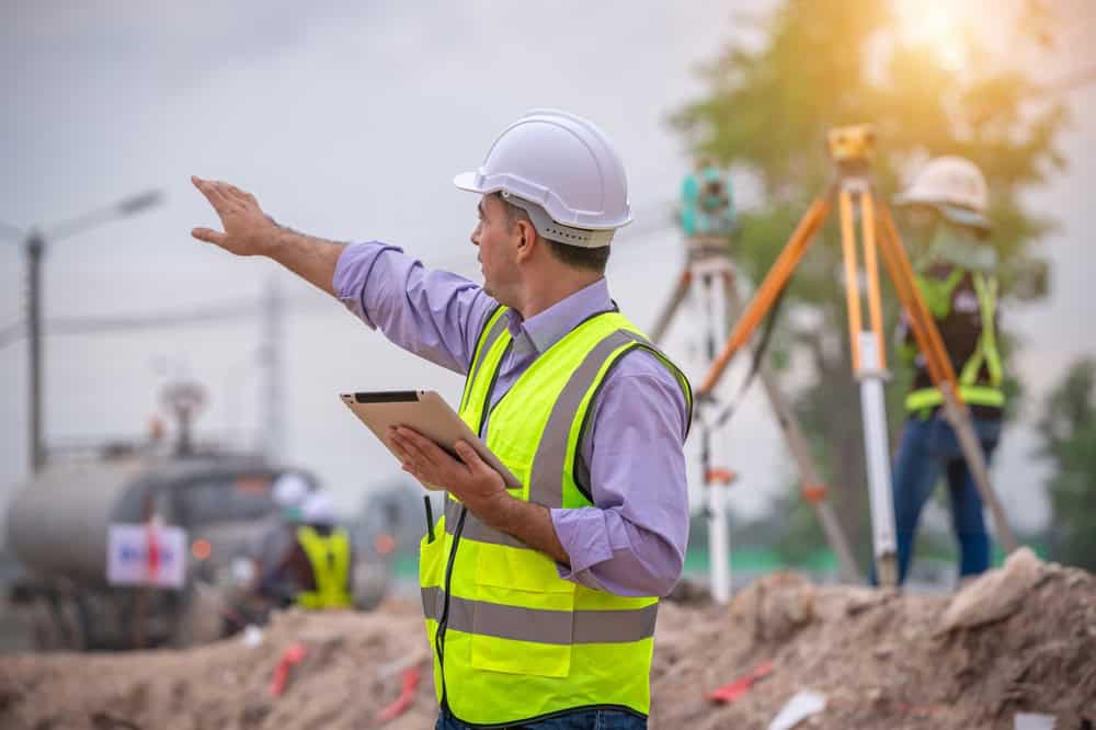 A construction worker in a reflective vest and hard hat holds a tablet, gesturing at a bustling construction site. Another worker operates surveying equipment in the background, exemplifying the precision of Land Surveyors in Okeechobee County. The scene is outdoors with machinery and trees visible.