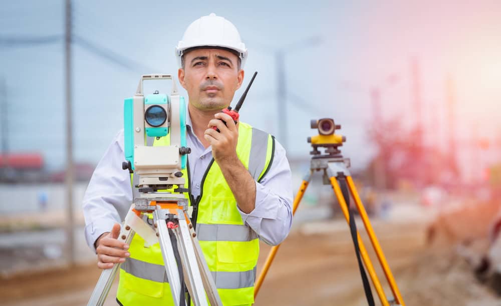 A construction worker in a white hard hat and yellow safety vest holds a walkie-talkie beside surveying equipment at a busy site. Land Surveyors Martin County might be involved, with another tripod visible in the background.