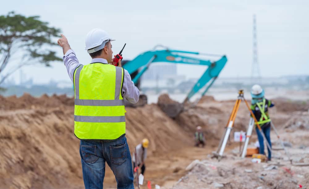 A construction site with a worker in a yellow reflective vest and hard hat using a walkie-talkie, pointing at an excavator. Land surveyors from Okeechobee County are surveying and digging in the background. A tree and power line tower are visible in the distance.