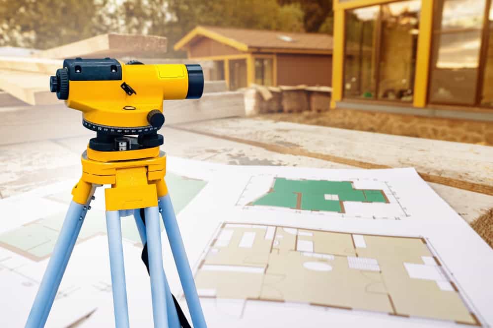 A yellow surveying instrument on a tripod stands on a construction site in Okeechobee County, with blueprints in the foreground. The blurred background includes wooden structures and walls under construction, showcasing the precision of land surveyors at work.