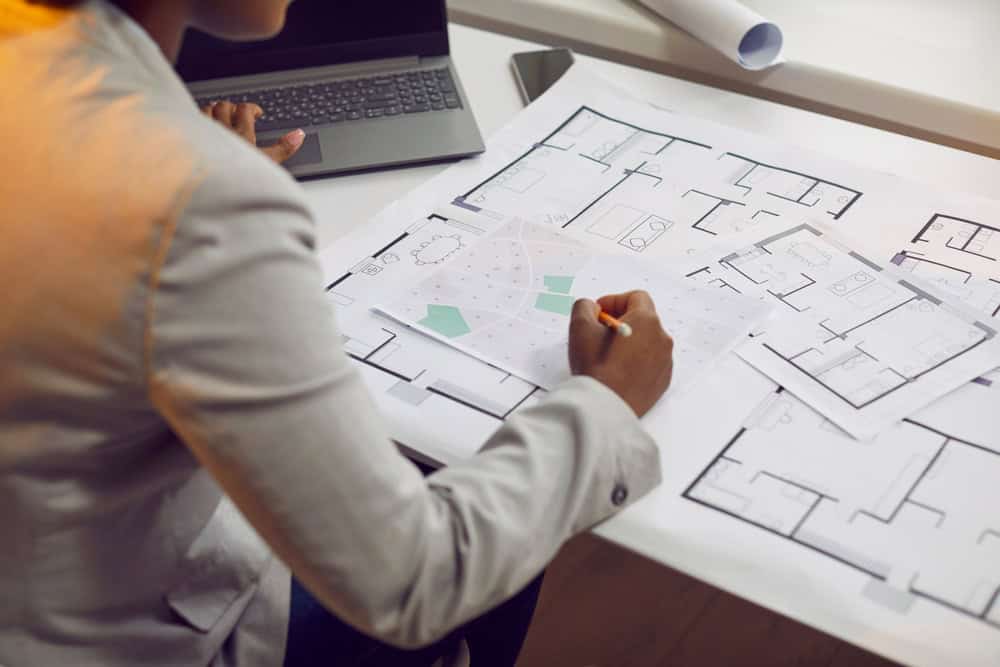 A person in a gray blazer is sitting at a desk, working on architectural blueprints with a pencil. A laptop is partially visible next to them while several detailed floor plans of Land Surveyors Martin County are spread out on the desk.