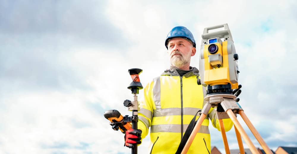 A surveyor wearing a blue hard hat and high-visibility jacket stands outdoors amidst the cloudy backdrop, holding equipment next to a tripod-mounted instrument, representing the precision and expertise of Land Surveyors in Indian River County.