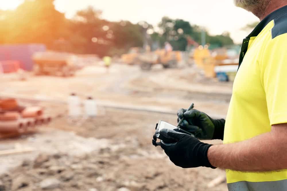 A construction worker in a yellow safety vest and black gloves writes on a notepad while standing on a bustling site, reminiscent of the precision seen with Land Surveyors in Martin County. The background reveals blurred machinery and materials under a bright sky.