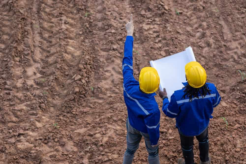 Two construction workers in blue jackets and yellow hard hats stand on a dirt field. One holds a blueprint, while the other points ahead, possibly discussing plans like seasoned land surveyors of Martin County. The ground shows parallel lines of soil preparation.