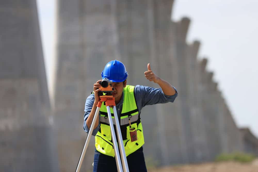 A construction worker in a blue hard hat and high-visibility vest confidently uses a theodolite for surveying, giving a thumbs-up gesture. Concrete structures form the backdrop, symbolizing the precision that land surveyors in Indian River County are known for.