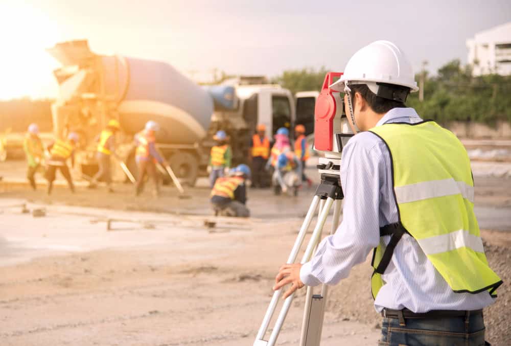 A construction worker wearing a safety vest and helmet uses surveying equipment at a busy site in Okeechobee County. In the background, other workers in similar attire work near a cement mixer. The scene is brightened by sunlight.